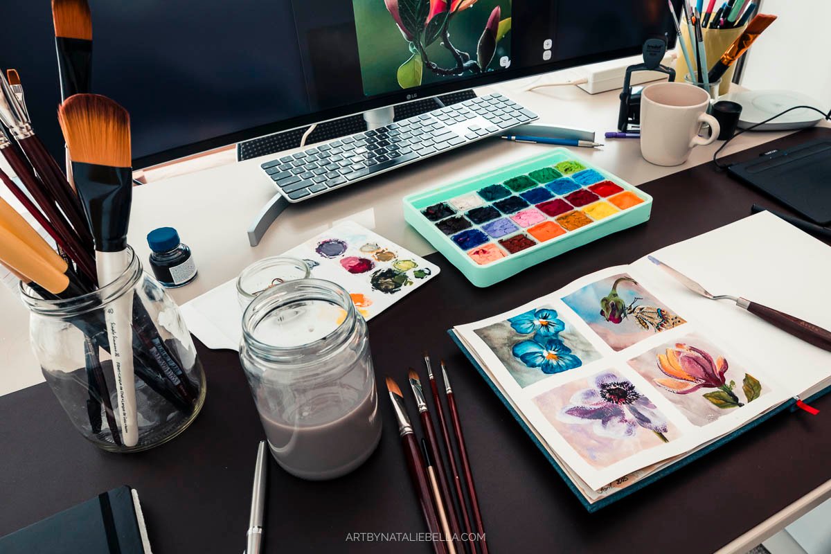 Artist workspace showing gouache paint palette, watercolor brushes in jar, open sketchbook with flower paintings, and computer on wooden desk