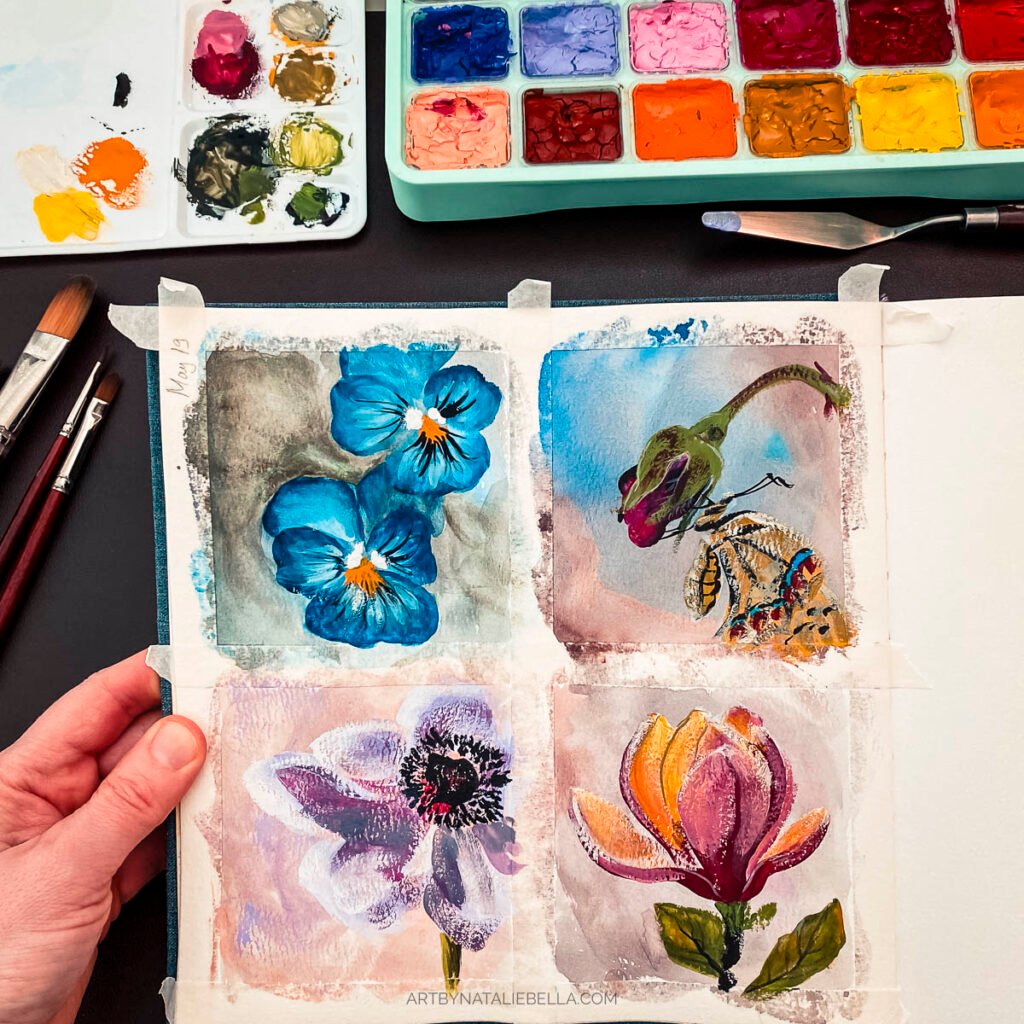 Artist's hand holding watercolor sketchbook with four gouache flower paintings, surrounded by colorful paint palette, mixing tray, and brushes on dark surface
