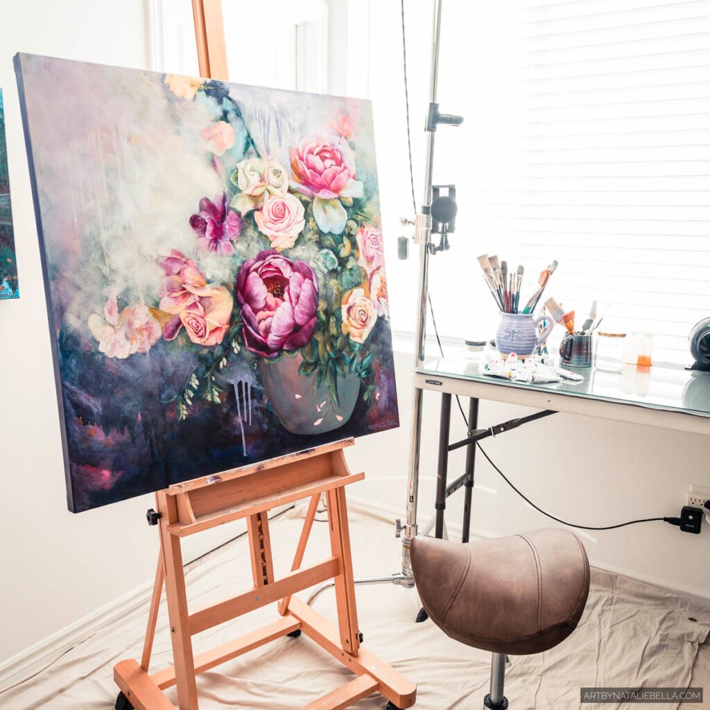 Mixed media painting "Floral Symphony" by Natalie Bella featuring vibrant pink peonies and soft roses displayed on a wooden easel in the artist's bright studio workspace, with art supplies, brushes, and natural lighting creating an authentic creative environment.