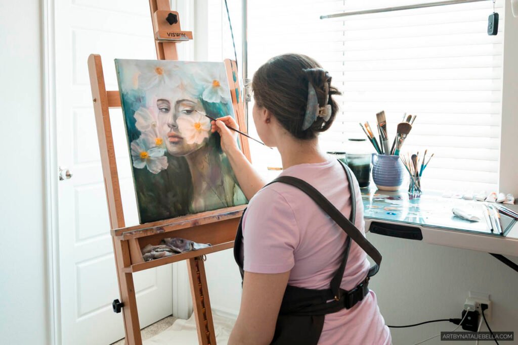 Female artist in pink shirt painting delicate portrait with white daisies on canvas in bright natural light studio with easel and brushes