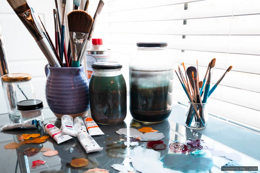 Glass artist palette with mixed oil paints, ceramic brush holders, paint tubes and brushes on bright studio table by window