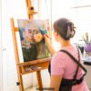 Female artist in pink shirt painting delicate portrait with white daisies on canvas in bright natural light studio with easel and brushes