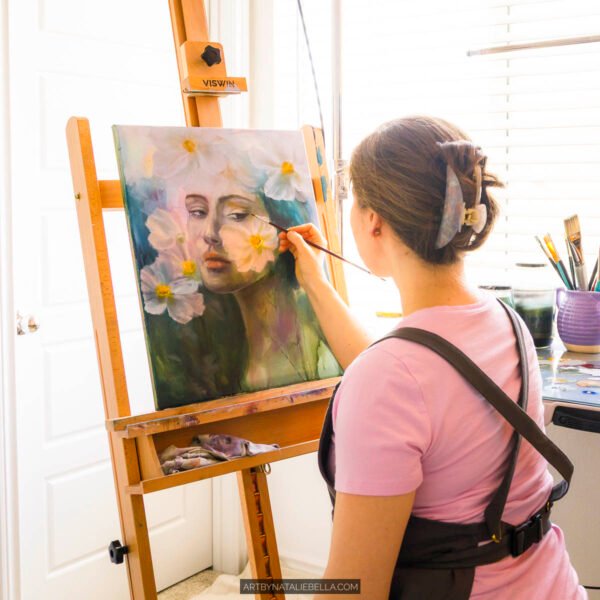 Female artist in pink shirt painting delicate portrait with white daisies on canvas in bright natural light studio with easel and brushes