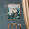 Where Ethereal Dreams Bloom original oil painting by Natalie Bella in walnut float frame displayed on textured blue-gray wall above vintage wooden chair with art books and white planter, showing ethereal woman portrait with white flowers in rustic home decor setting