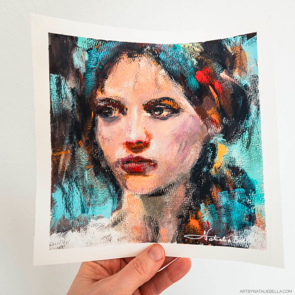 A hand holds the completed gouache painting 'Unguarded' against a white background, showing the full 8x8 inch artwork on watercolor paper. The expressive portrait of a woman features bold brushstrokes in turquoise, orange, and warm skin tones, with her direct gaze and red lips as focal points. The artist's signature is visible in white in the bottom right corner. The hand holding the piece gives scale to the intimate size of this vibrant, textural portrait.