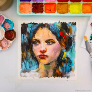 Studio flat lay showing the gouache painting 'Unguarded' - an 8x8 inch portrait of a woman with expressive eyes and red lips, painted in vibrant blues, oranges, and warm skin tones on watercolor paper. The painting is surrounded by art supplies including a well-used watercolor palette with colorful paint wells, a palette knife, and small containers of mixed paint, all arranged on a white surface.