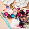 An artist's workspace featuring white ceramic flower-shaped paint palettes filled with vibrant gouache colors, mint green paintbrushes, and a colorful abstract portrait painting on paper with expressive brushstrokes in purples, magentas, and earth tones, all arranged on a light wood surface