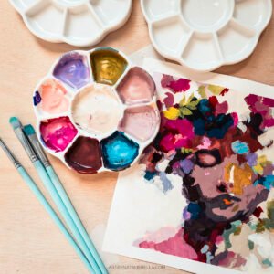 An artist's workspace featuring white ceramic flower-shaped paint palettes filled with vibrant gouache colors, mint green paintbrushes, and a colorful abstract portrait painting on paper with expressive brushstrokes in purples, magentas, and earth tones, all arranged on a light wood surface
