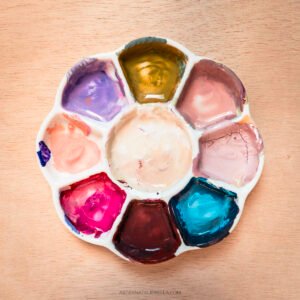 A white ceramic flower-shaped paint palette with eight petals containing various gouache paints in vibrant colors including purple, golden yellow, coral pink, magenta, deep brown, turquoise blue, and soft pink tones, sitting on a light wood surface