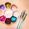 A white ceramic flower-shaped paint palette filled with vibrant gouache paints in purple, golden yellow, coral pink, magenta, deep brown, and turquoise blue, alongside three mint green artist brushes with silver ferrules on a light wood surface