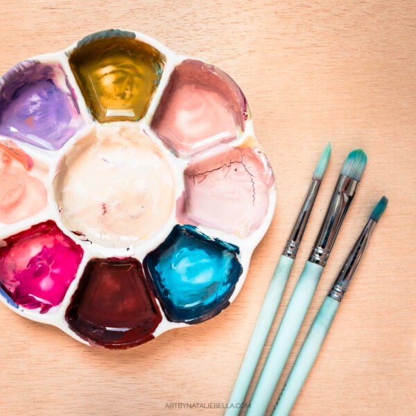 A white ceramic flower-shaped paint palette filled with vibrant gouache paints in purple, golden yellow, coral pink, magenta, deep brown, and turquoise blue, alongside three mint green artist brushes with silver ferrules on a light wood surface