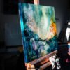 Awakening - Teal Abstract Painting on Easel in Artist Studio by Original 24x24 teal abstract painting Awakening displayed on an easel in the Las Vegas studio of artist Natalie Bella, with other artworks visible in the background