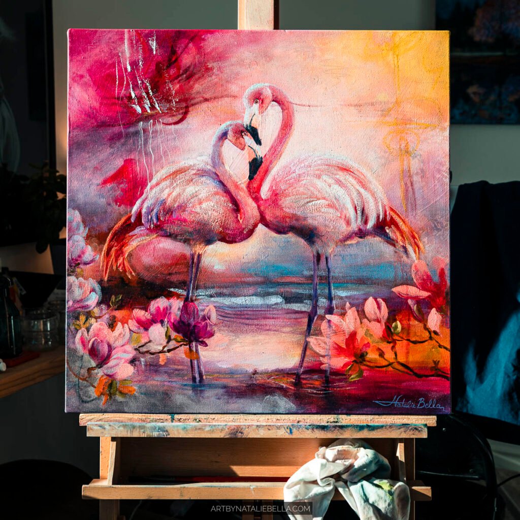 Coastal Grace, 24x24 inch oil and mixed media painting by Natalie Bella displayed on wooden easel showing two flamingos in tropical setting with pink florals